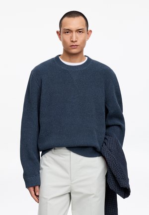 Mann trägt dunkelblauen Strickpullover und hellbeige Hose, hält eine marineblaue karierte Jacke in seiner linken Hand und steht vor einem weißen Hintergrund.