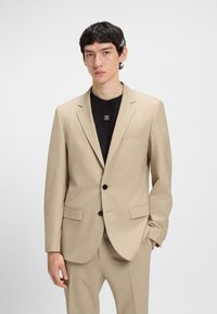 Young man with short dark hair wearing a beige two-button suit and black shirt with "HUGO" logo on a plain white background.