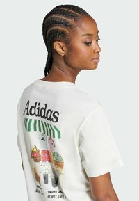 Cream cotton t-shirt featuring "Adidas" text on the back, with colorful graphics of clothing and potted plants. Short sleeves and crew neck design.