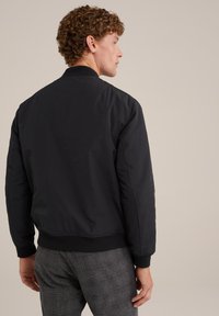 Černá bomber bunda s hladkou texturou, žebrovanými manžetami a pasem, spárovaná s strukturovanými šedými kostkovanými kalhotami, viděná zezadu.