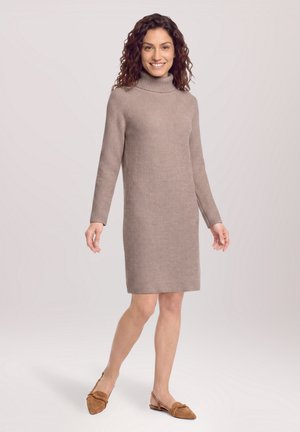 Femme aux cheveux bouclés portant une robe en maille beige à col roulé et des chaussures plates marron, debout et souriante sur un fond clair uni.