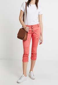 Camiseta blanca ligera, pantalones capri color coral con detalles de botón y cremallera, y un bolso cruzado marrón. Zapatillas deportivas blancas completan el look.