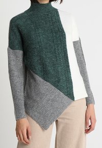 Pull en tricot texturé avec un col haut, comportant des sections diagonales en vert foncé, blanc et gris. Ourlet asymétrique et manches côtelées.