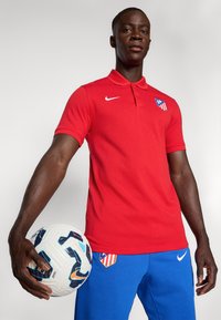 Rode polo met kraag en een wit Nike-logo, gecombineerd met blauwe broek met een logo-patch. Het model houdt een witte voetbalschoen vast.
