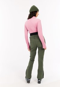 Vrouw in een roze lange mouwtop, olijfgroene broek, groene beanie en zwarte skischolen, staande met haar rug naar de camera op een witte achtergrond.