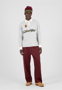 Šedý polo s dlhým rukávom a bielym golierom s logom "Social Club"; skombinovaný s bordovými nohavicami s širokými nohavicami a béžovými topánkami. Na hlave burgundská čiapka.