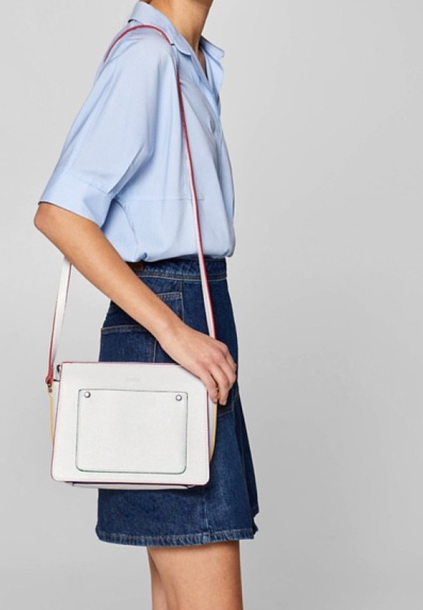 Bolso rectangular blanco con un bolsillo frontal transparente, ribete negro y una correa ajustable colorida, usado cruzado. Material texturizado.
