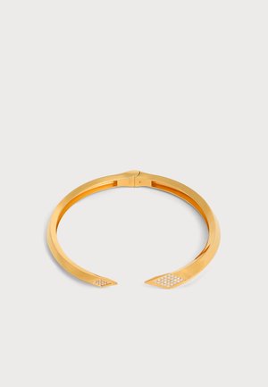 Bracelet manchette ouvert en or avec des extrémités pointues, chacune décorée de petites pierres blanches, sur un fond clair uni.