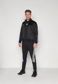 Fato de treino atlético preto com uma jaqueta com fecho éclair e calças combinando. Apresenta detalhes de três riscas brancas e logótipo na jaqueta.