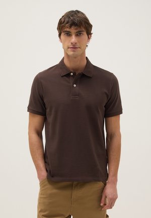 Joven con camiseta polo de manga corta marrón y pantalones color caqui, de pie con una mano en el bolsillo contra un fondo liso.