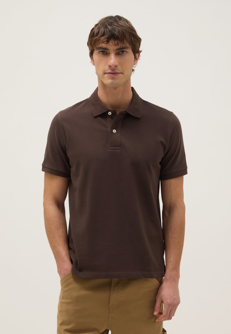 Joven con camiseta polo de manga corta marrón y pantalones color caqui, de pie con una mano en el bolsillo contra un fondo liso.