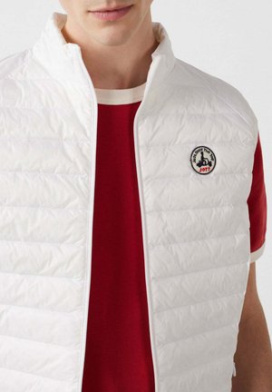 Giovane che indossa un gilet bianco trapuntato sopra una maglietta a maniche corte rossa e bianca, con una toppa rotonda con logo sul lato sinistro del petto del gilet.