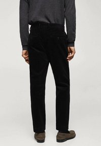 Homme portant un pantalon en velours côtelé noir, une chemise à manches longues grise et des mocassins marron, debout de dos sur un fond uni.