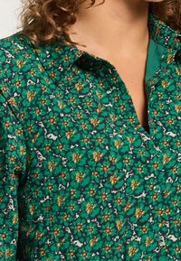 Grön blommig skjorta med bred krage, små knappar och en texturerad tyg med gula och mörkgröna blommönster.