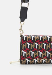 Torebka typu crossbody Tommy Hilfiger z geometrycznym wzorem w kolorze czerwonym, białym i złotym na czarnym tle; posiada złoty zamek błyskawiczny i odpinany pasek.