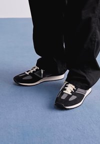 Person wearing black and gray sneakers with white laces, standing on a light blue carpet, dressed in black pants.