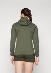 Grønn, langermet hettegenser med strukturert overflate, grå detaljer på skuldre og mansjetter, samt en figursydd silhuett, brukt over mørke shorts.
