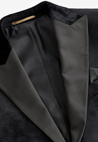 Black tuxedo jacket featuring a sleek satin lapel, textured fabric interior, and a pocket square. Smooth and soft materials throughout.