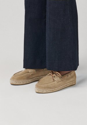 Mocassins espadrilles en daim beige avec semelles en corde, détails à lacets, portés avec un pantalon en denim large et foncé.
