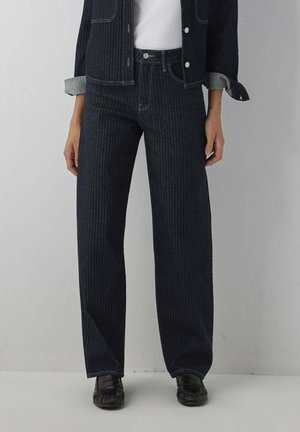 Person trägt dunkle, dunkel gestreifte Jeans mit geradem Bein, passende Jacke mit hochgekrempelten Ärmeln, weißes Top und schwarze Loafers, steht vor einer schlichten Wand.
