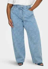 Jean large couleur bleu clair avec un motif floral subtil, taille avec cordon de serrage, poches avant, porté avec des talons ouverts et un haut blanc.