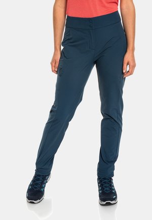 Femme portant un pantalon d'extérieur bleu marine avec des poches à fermeture éclair à l'avant et sur les côtés, assorti à des chaussures de randonnée noires et bleues et un haut corail.