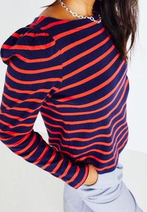 Femme portant un pull bleu marine à rayures horizontales rouges et épaules bouffantes, associé à un pantalon gris clair et un collier chaîne argenté.