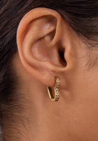 Goldene Creolen mit einem wellenförmigen Design, verziert mit kleinen, klaren Edelsteinen auf einer Seite; abgebildet am Ohr vor einem neutralen Hintergrund.