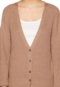 Frau, die eine gerippte, geknöpfte, hellbraune Strickjacke über einem weißen Top mit Rundhalsausschnitt trägt, dargestellt von den Schultern bis zur Mitte des Oberkörpers.