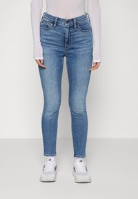 Jeans de ganga azul de cintura alta, com um corte justo, design de cinco bolsos e detalhes cosidos. Usados com uma camisa de manga longa branca e ténis.