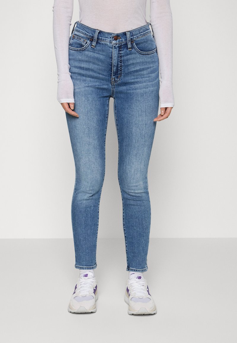 Jeans de ganga azul de cintura alta, com um corte justo, design de cinco bolsos e detalhes cosidos. Usados com uma camisa de manga longa branca e ténis.