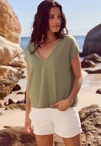 Top de punto con escote en V en suave verde oliva con tela texturizada, combinado con pantalones cortos blancos remangados. Escenario de playa con rocas y agua de fondo.