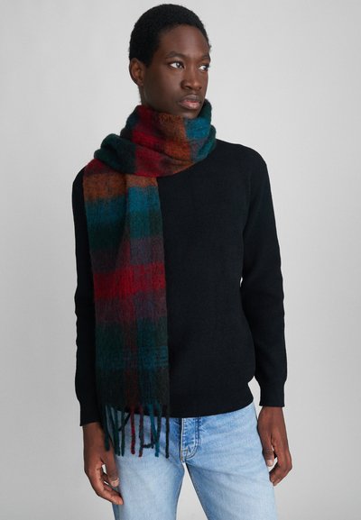 Giovane uomo che indossa un maglione nero, jeans azzurri, e una sciarpa a quadri rossi, blu e verdi, che guarda di lato su uno sfondo semplice.