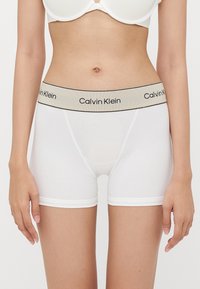 Bijele pamučne bokserice s bež pojasom na kojem je crni branding "Calvin Klein". Pokazuju gladak materijal i prilagođeni kroj.