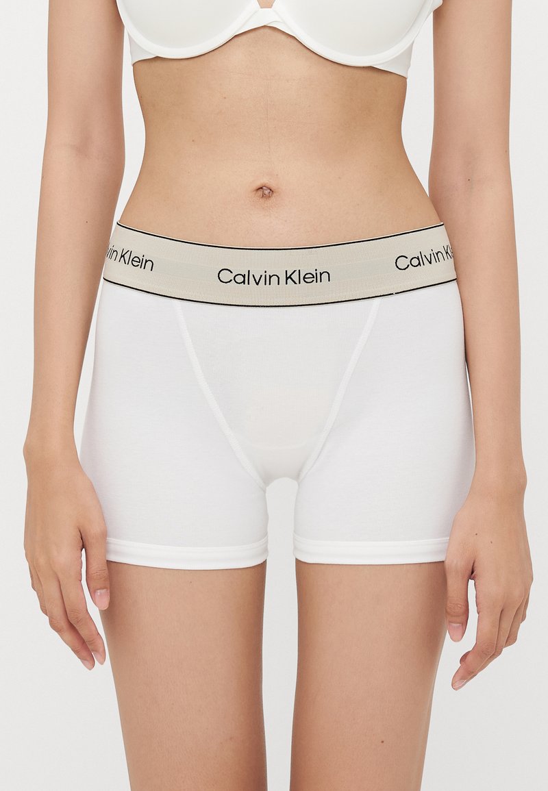 Bijele pamučne bokserice s bež pojasom na kojem je crni branding "Calvin Klein". Pokazuju gladak materijal i prilagođeni kroj.