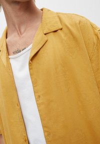 Männlicher Oberkörper, der ein senfgelbes gemustertes Hemd über einem weißen Tanktop trägt, und ein Brusttattoo zeigt, das "vida" lautet.