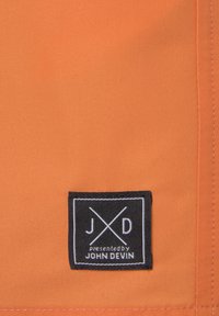 Narančasta tkanina glatke teksture sadrži crnu oznaku na kojoj piše "JXD" i "Presented by JOHN DEVIN" u bijelom tekstu, s križnim linijama.