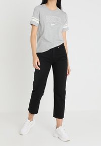 Mujer vistiendo una camiseta gris de Nike con rayas blancas en las mangas, pantalones cortos negros y zapatillas blancas, de pie contra un fondo liso.