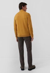 Muž v horčicovo žltom svetri, tmavo sivých nohaviciach a hnedých topánkach, stojí čelom k obyčajnej svetlo sivej stene.