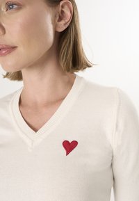 Romantische V-hals trui in crèmekleurige stof met een zachte textuur, voorzien van een klein rood geborduurd hartje op de linkerborst.