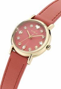 Reloj con marco dorado, esfera rosa, marcadores de hora de cristal que incluyen una forma de corazón y una correa de cuero rosa.
