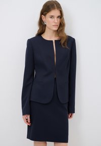 Tummansininen räätälöity bleiseri, jossa on etuhalkio, pitkät hihat ja kaksi taskua, yhdistettynä samasta kankaasta valmistettuun yksilölliseen hameeseen. Teksturoitu kangas.