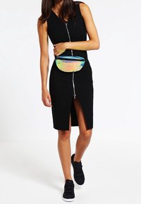 Abito nero senza maniche con zip frontale, spacco fino al ginocchio e una borsa a tracolla olografica colorata; abbinato a sneakers nere con velcro.