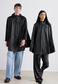 To modeller har matchende svarte hetteponchoer i et glatt, blankt stoff. Ponchoene har en avslappet passform og er kombinert med løse blå jeans.