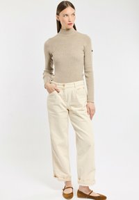 Une femme se tient debout, portant un col roulé beige à côtes, un pantalon à large jambe blanc cassé avec des revers, des chaussures plates marron, les mains dans les poches, regardant sur le côté.