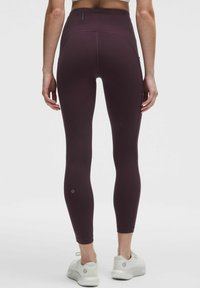 Temno vijolične hlače z visokim pasom, izdelane iz raztegljivega, teksturiranega materiala, s sleek oblikovanjem in logotipom blizu gležnja, v kombinaciji z belimi čevlji.