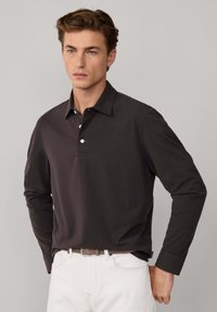 Polo a maniche lunghe di colore marrone scuro, con colletto, patta con tre bottoni e superficie liscia. Indossato con pantaloni chiari.