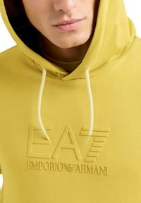 Uomo che indossa una felpa con cappuccio gialla con il logo in rilievo "EA7 Emporio Armani" e i lacci visibili sul cappuccio.