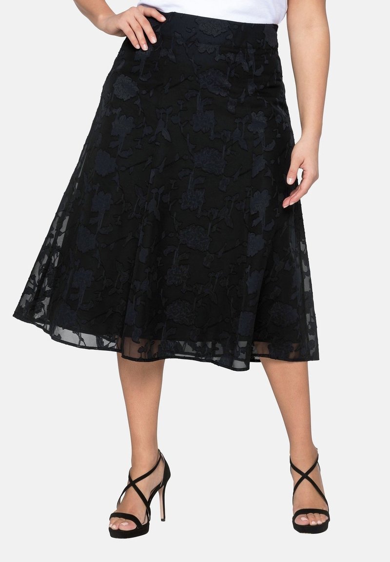 Falda negra acampanada con superposición de encaje floral, combinada con sandalias de tacón alto negras y una blusa blanca, mostrada de la cintura a los pies.