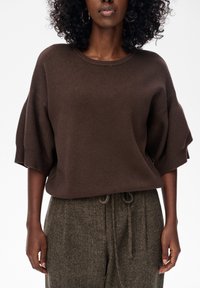 Vrouw in een losse bruine gebreide trui met ellebooglange mouwen en een hoge taille met textuur in de broek en een tailleband met trekkoord.